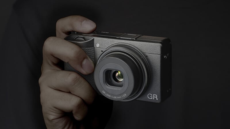 A hand holding a compact digital camera with the lens extended against a dark background. The camera has "GR" printed on the front.