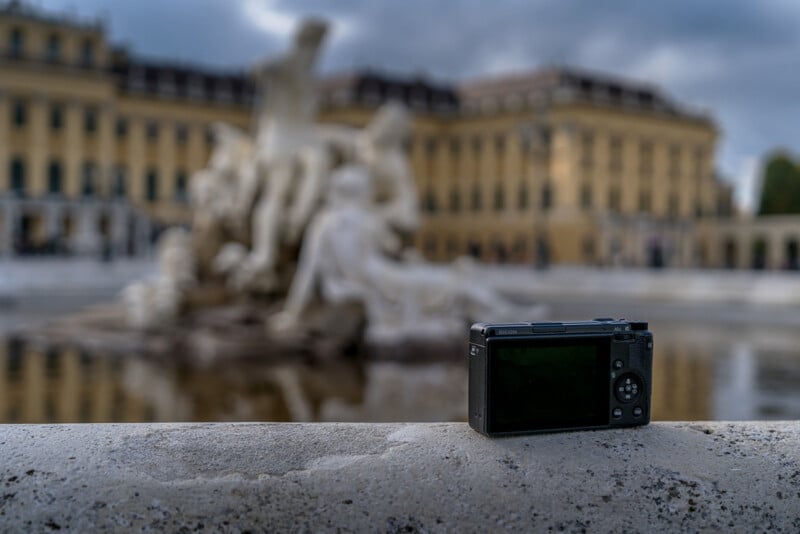 A digital camera rests on a stone ledge in front of a blurred fountain and historic yellow building, possibly a palace, under a cloudy sky.