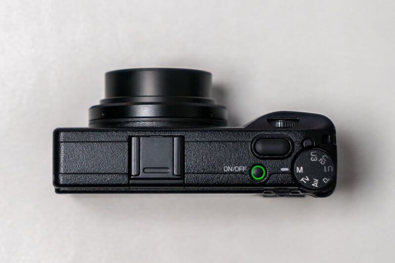 Top view of a compact black digital camera with a partially extended lens, mode dial, on/off button with a green ring, and a textured surface, placed on a light background.