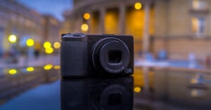 A black compact digital camera is placed on a reflective surface outdoors, with a blurred background of city buildings and circular yellow lights.