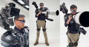 A man wearing sunglasses and a futuristic black and gray exoskeleton suit holds and operates a large camera with an oversized telephoto lens in a white studio setting. The suit has mounted camera gear on his shoulders.
