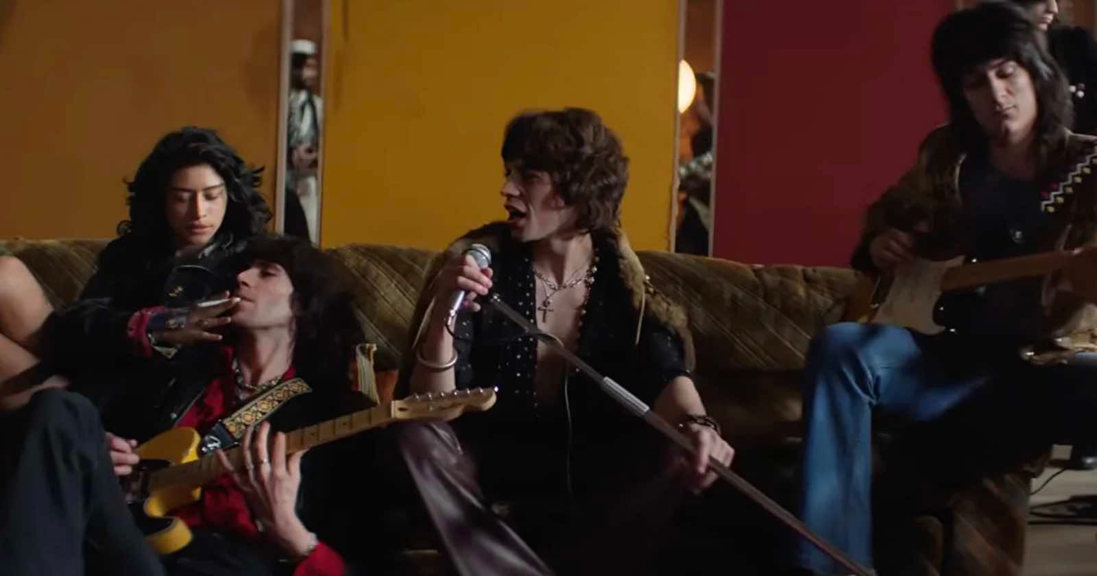 Four people sit on a couch; one sings into a microphone, two play electric guitars, and one reclines while another holds their hand. The background features orange and red walls.