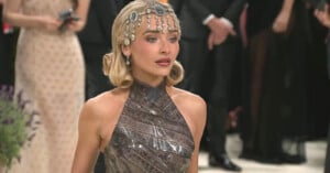 A woman wearing an ornate jeweled headpiece and a metallic, patterned halter dress stands in a formal setting, with people dressed in evening attire blurred in the background.