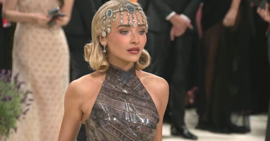 A woman wearing an ornate jeweled headpiece and a metallic, patterned halter dress stands in a formal setting, with people dressed in evening attire blurred in the background.