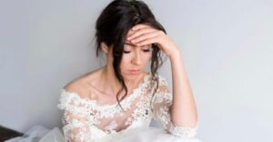 A woman in a white lace wedding dress sits with her head resting on her hand, looking down with a sad or thoughtful expression against a plain light background.