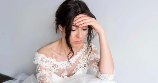 A woman in a white lace wedding dress sits with her head resting on her hand, looking down with a sad or thoughtful expression against a plain light background.