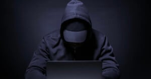 A person in a dark hoodie and cap sits in front of a laptop, their face obscured by shadows, creating a mysterious and anonymous atmosphere.