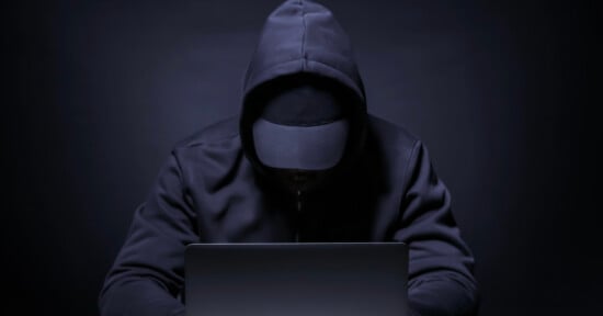 A person in a dark hoodie and cap sits in front of a laptop, their face obscured by shadows, creating a mysterious and anonymous atmosphere.