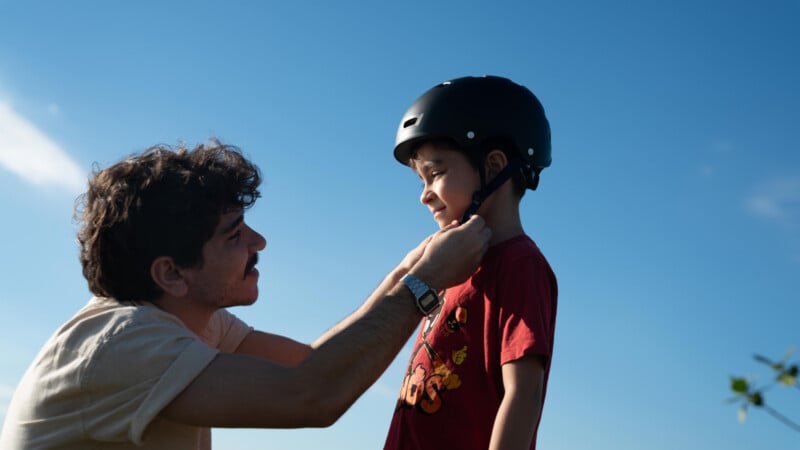 A man kneels to fasten a black helmet on a smiling child wearing a red shirt, with a clear blue sky in the background.