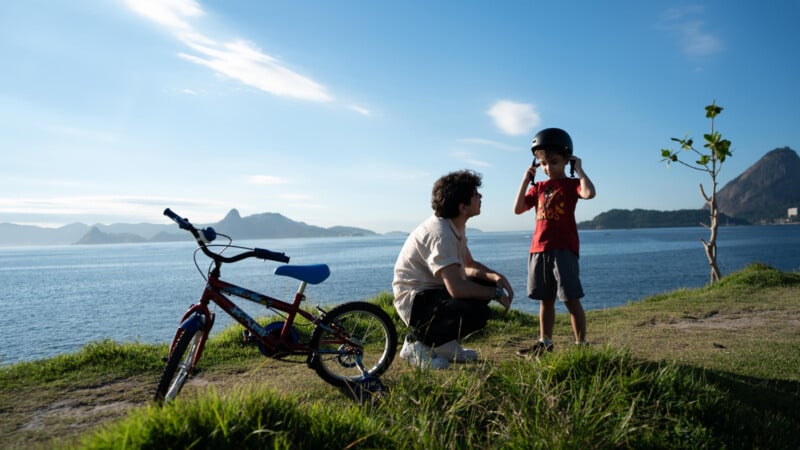 An adult crouches beside a child adjusting a helmet near a bicycle on a grassy hill overlooking a large body of water, with distant mountains and a clear blue sky in the background.