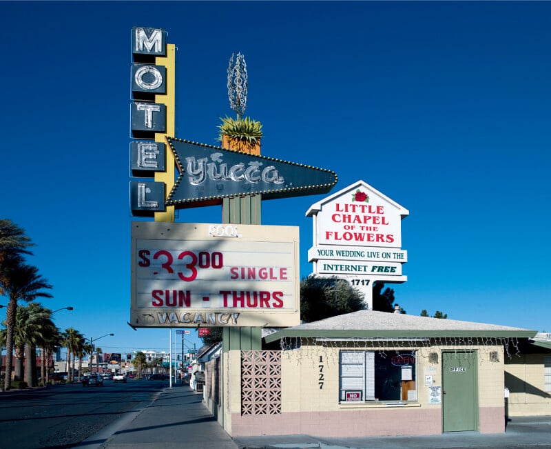 A vintage "Motel Yucca" sign displays rates and vacancy under a clear blue sky. Next to it, a sign for "Little Chapel of the Flowers" advertises free internet wedding viewing. Palm trees and a small building are visible.
