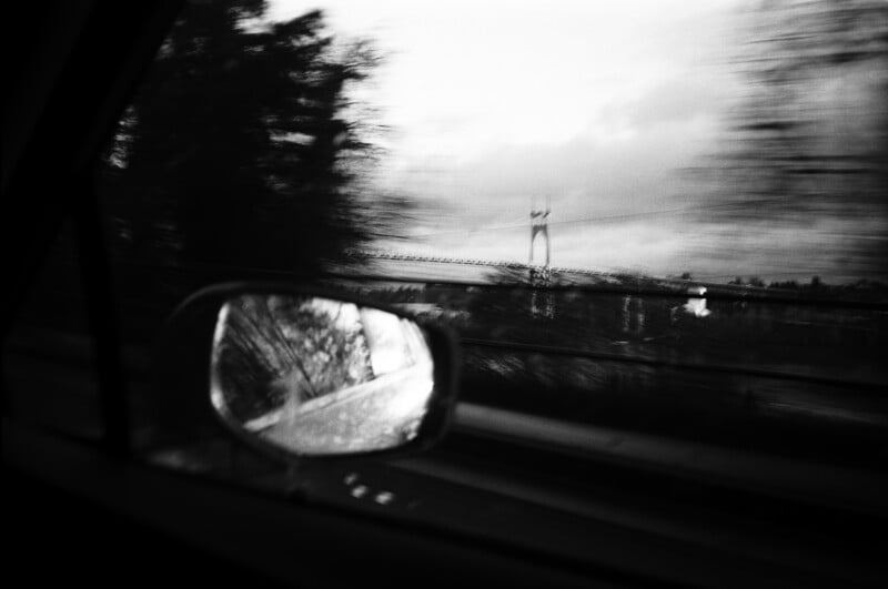 Black-and-white photo taken from inside a moving car, showing a blurry landscape with trees and a bridge outside. The car’s side mirror reflects part of the scene. The image has a sense of motion and speed.