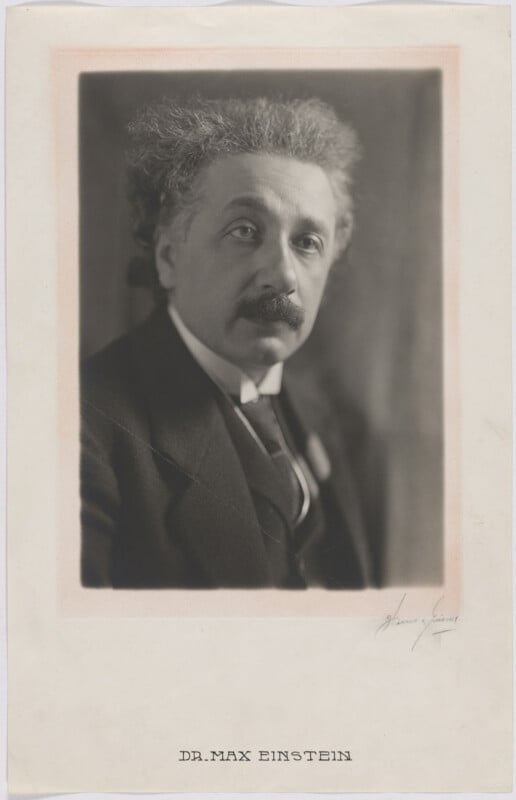 A black-and-white portrait of a man with wild, curly hair and a mustache, wearing a suit and tie. The photograph is mounted on paper labeled "DR. MAX EINSTEIN.