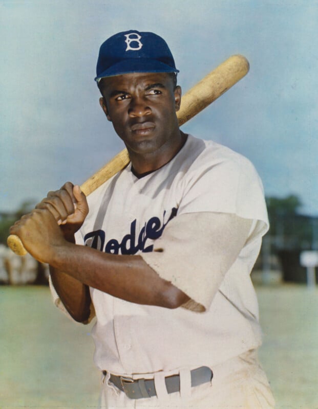 A baseball player in a Brooklyn Dodgers uniform stands holding a bat on his shoulder, looking to the side. The background is an outdoor field with a blue sky.