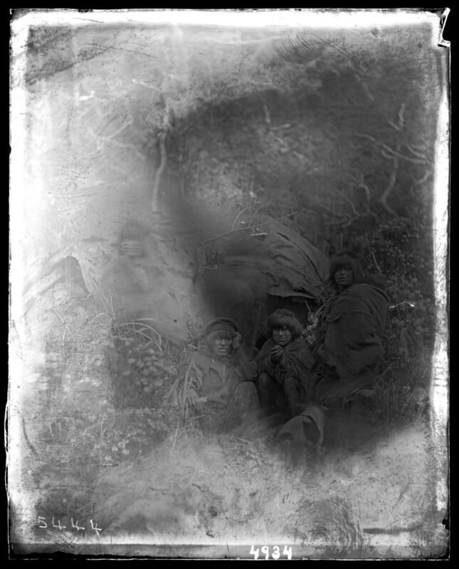 A black-and-white, partially faded photo shows four people sitting outdoors in a natural setting, wrapped in thick blankets or clothing, surrounded by foliage and trees. The image is blurry and weathered with visible damage.
