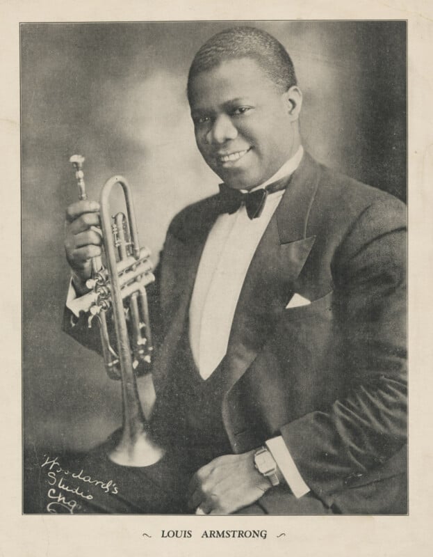 Black-and-white portrait of Louis Armstrong in a tuxedo, smiling and holding a trumpet upright; his signature appears on the bottom left of the photo.