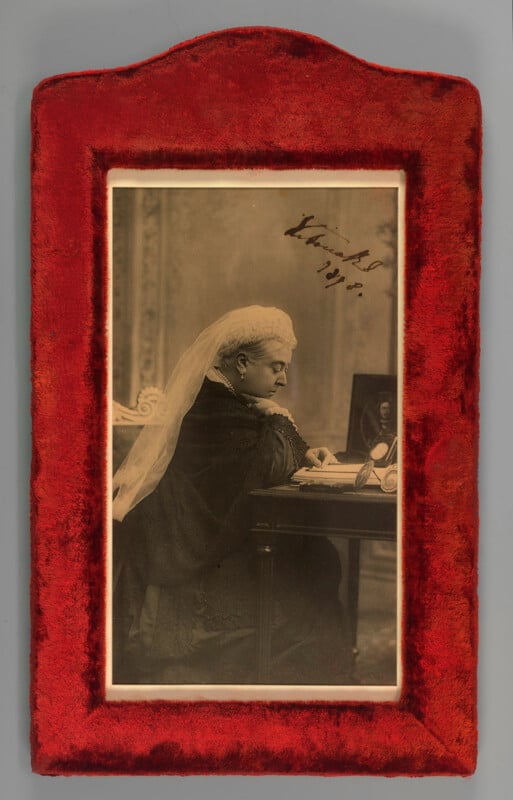 Sepia photograph in a red velvet frame shows a woman in profile, wearing dark clothing and a white veil, seated at a desk with papers and a framed photo. There is handwriting in ink on the upper right of the image.