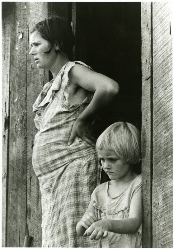 A pregnant woman in a plaid dress stands in a wooden doorway with her hand on her hip, looking out pensively. Beside her, a young girl with short blond hair stands, also gazing outside, appearing thoughtful.