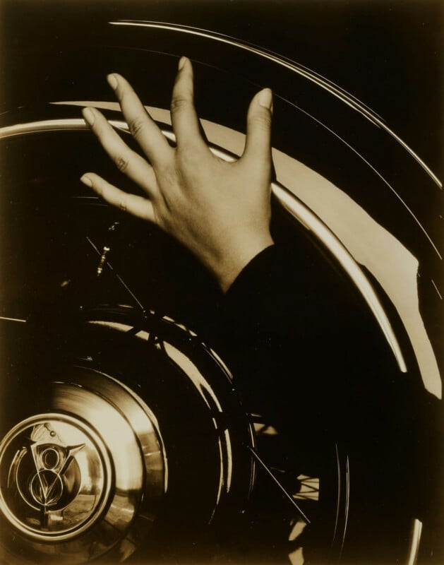 A hand with outstretched fingers rests on a shiny, reflective car wheel with an intricate hubcap. The image is in sepia tones, highlighting the contrast between the hand and the metallic surface.