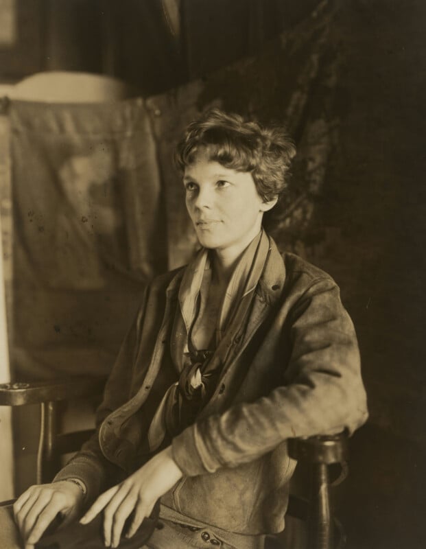 A sepia-toned portrait of a woman with short, wavy hair, wearing a jacket and scarf, seated on a wooden chair with one arm resting on the armrest. The background is blurred fabric or drapery.