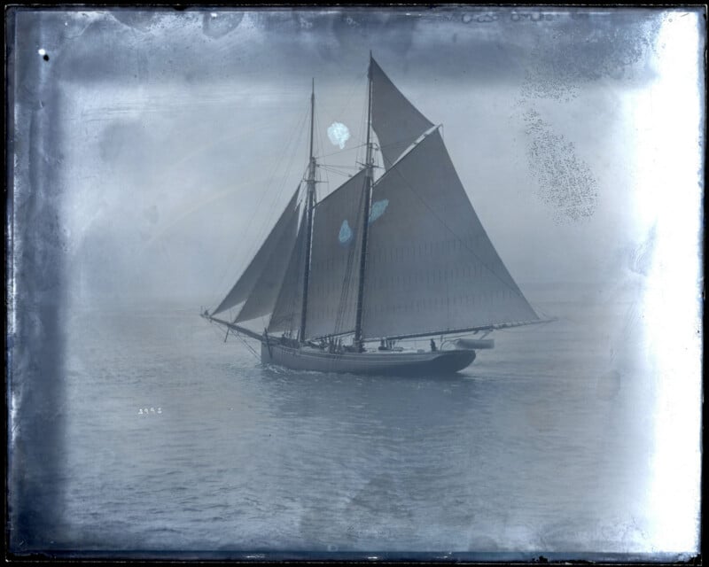 A vintage photograph of a large sailboat with two masts and dark sails gliding on calm water, surrounded by a misty or foggy atmosphere. The image has scratches and visible signs of age.