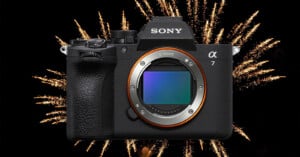 A Sony Alpha 7 camera is centered in the image with its lens removed, revealing the sensor, while bright fireworks explode in the black night sky behind it.