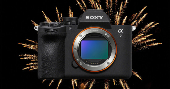 A Sony Alpha 7 camera is centered in the image with its lens removed, revealing the sensor, while bright fireworks explode in the black night sky behind it.