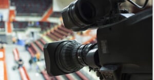 Close-up of a professional video camera focused on an indoor sports arena with empty red and beige seats visible in the blurred background.