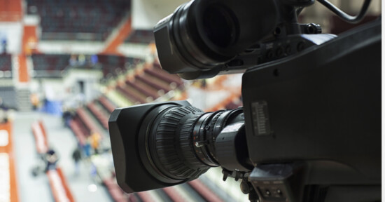 Close-up of a professional video camera focused on an indoor sports arena with empty red and beige seats visible in the blurred background.