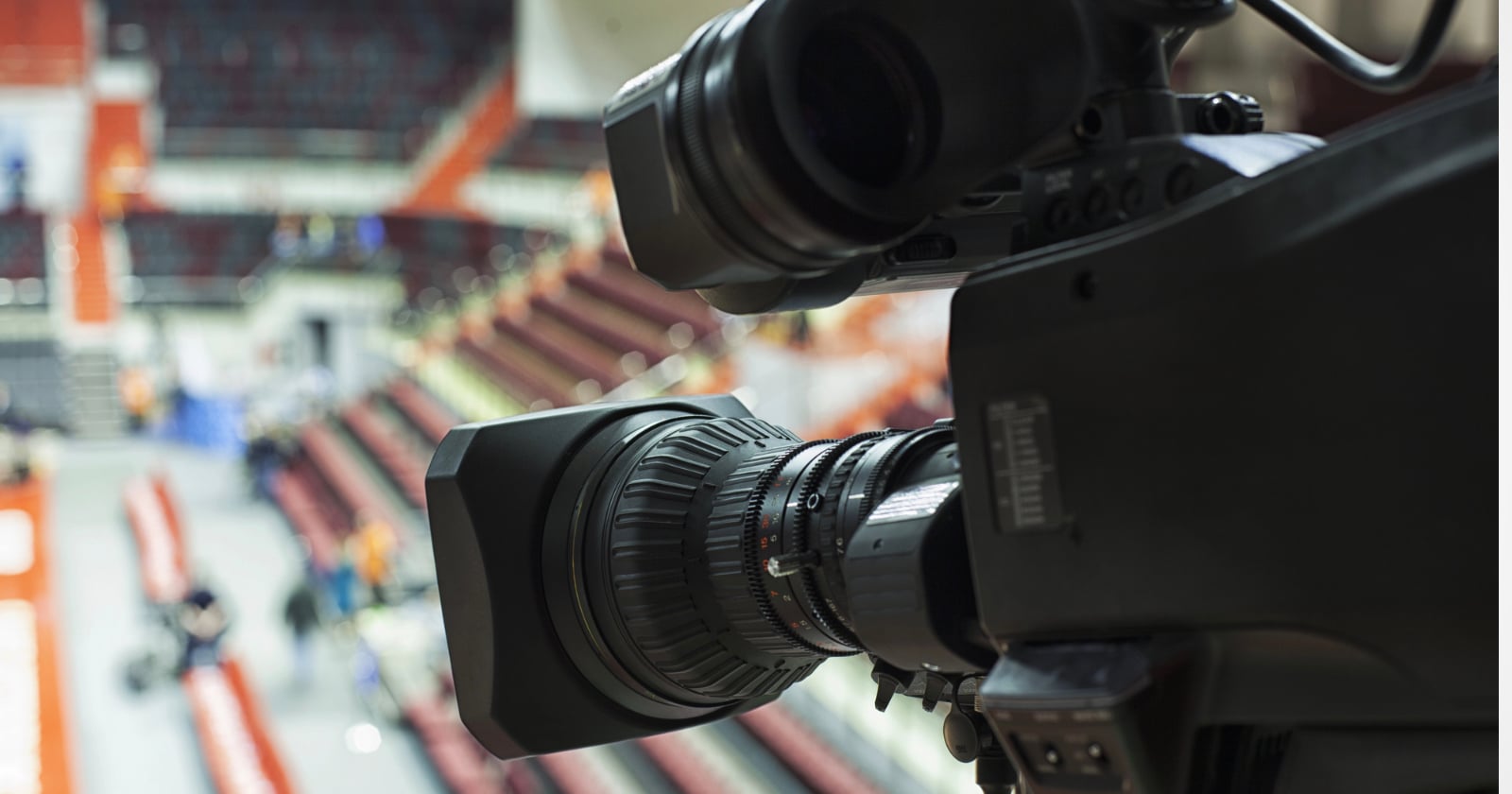 Close-up of a professional video camera focused on an indoor sports arena with empty red and beige seats visible in the blurred background.