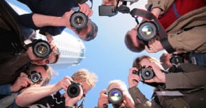 A group of photographers stands in a circle, aiming their cameras downward, capturing a shot from above on a sunny day with a tall building in the background and blue sky overhead.