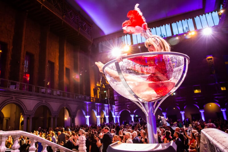 A performer in a red sequined outfit and feathered headdress poses inside a giant martini glass on stage, overlooking a large, elegantly dressed crowd in a grand hall with purple lighting.