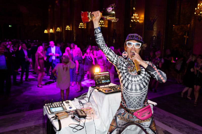 A lively performer in a patterned bodysuit, white glasses, and a pink fanny pack holds a trophy and microphone on a stage, with a dancing crowd and purple lights in the background.