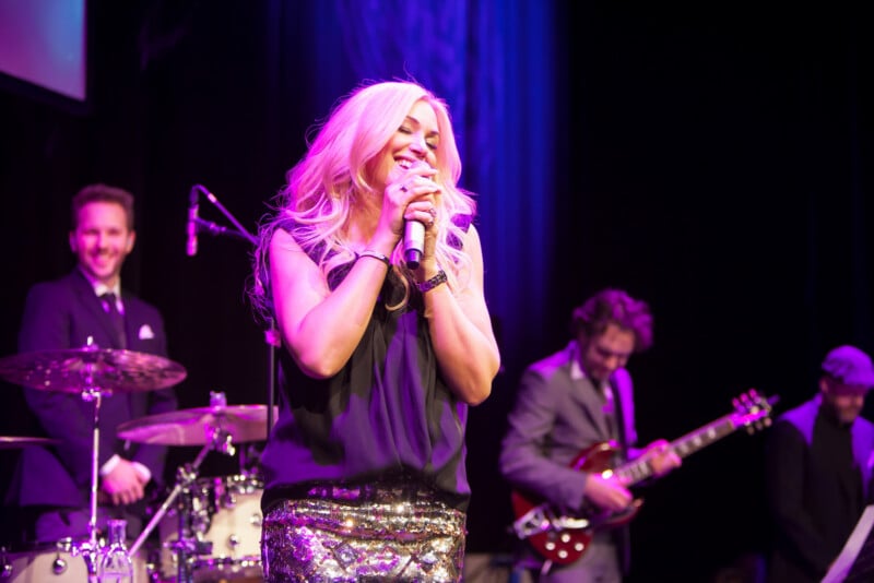 A blonde woman sings on stage, smiling with her eyes closed and holding a microphone. She wears a black top and a sparkling skirt. Behind her, three male musicians play drums, guitar, and keyboard under purple lighting.