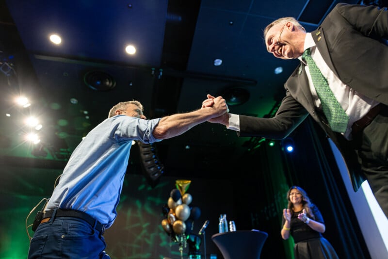 Two men clasp hands in a handshake on stage, one in a suit and the other in a shirt and jeans. A woman stands in the background clapping, with balloons and stage lights behind them.