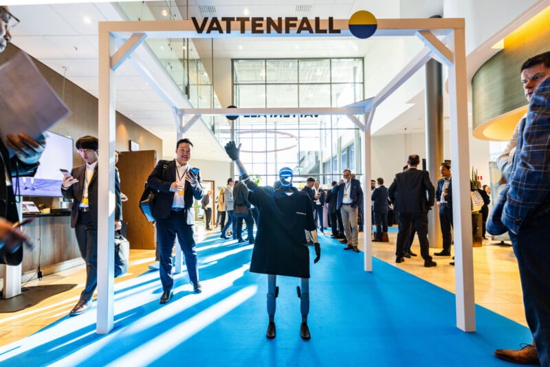 A humanoid robot stands under a white frame labeled "Vattenfall" at a busy indoor event, surrounded by people in business attire, some interacting and some using their phones.