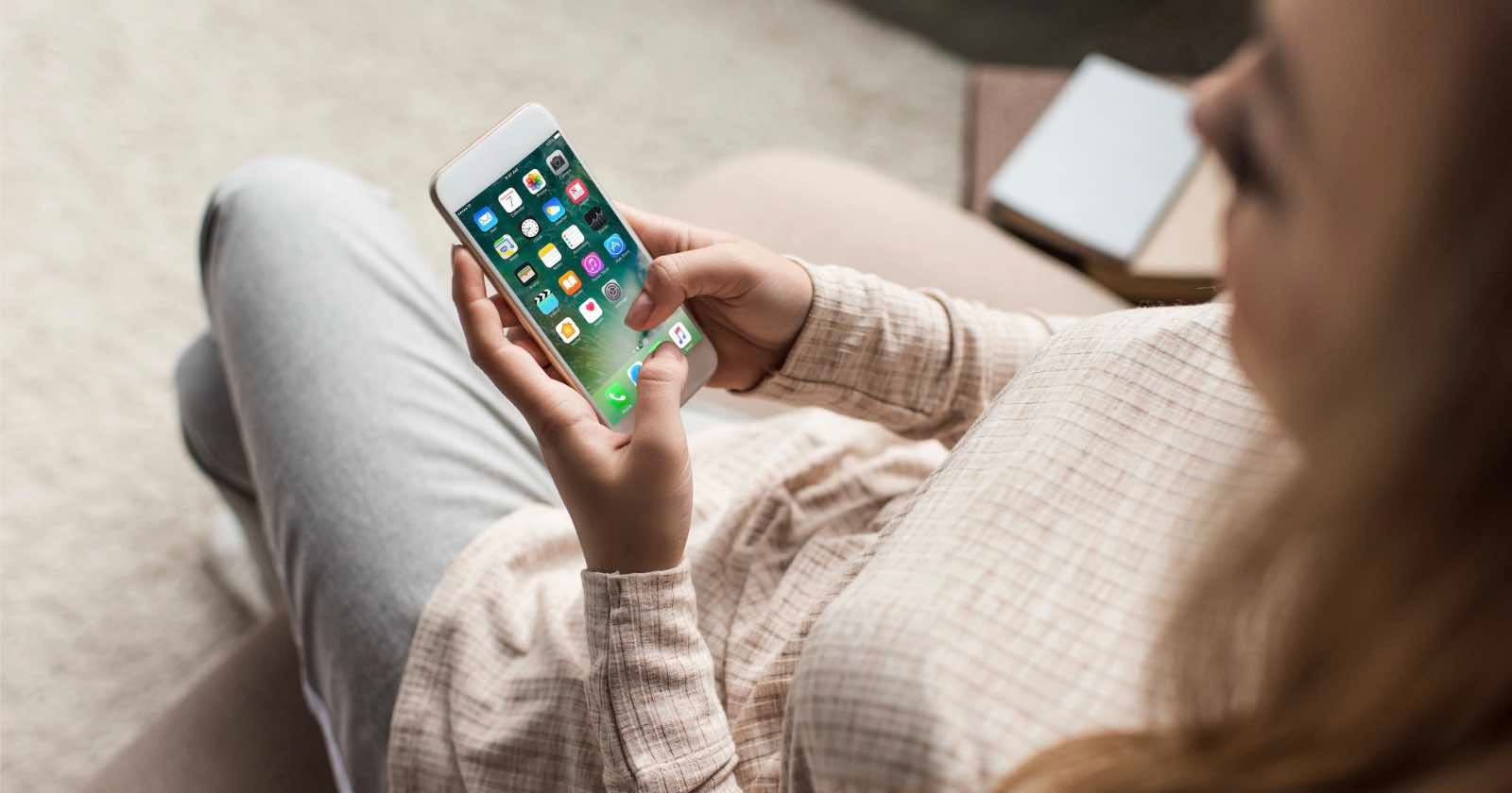 A person sitting on a couch uses a smartphone, holding it in both hands. The phone screen displays various app icons. The person is wearing light-colored clothes and is viewed from above.