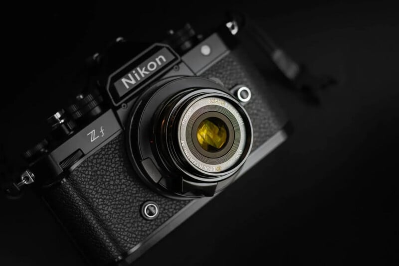 Close-up of a black Nikon Z f camera with a textured body and a lens reflecting yellow light, set against a dark, blurred background.