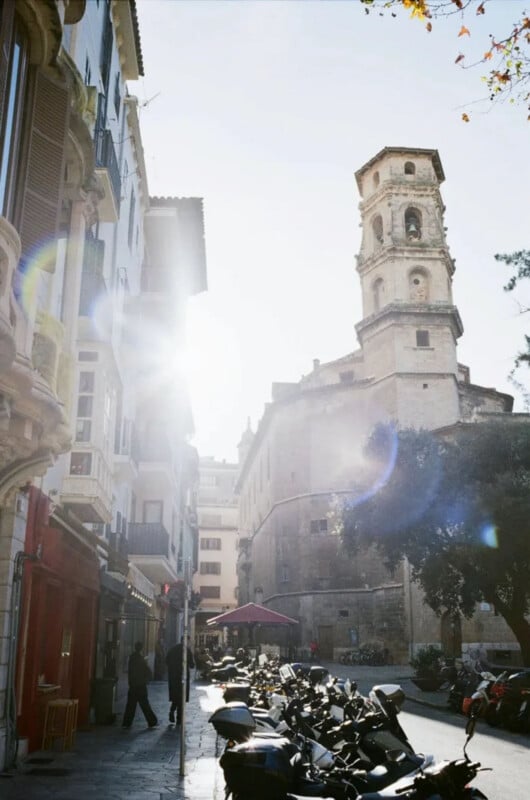 A sunlit street scene in a European city features a tall church tower, historic buildings, parked motorcycles, and people walking near an outdoor café. Sunlight creates lens flares and casts long shadows.
