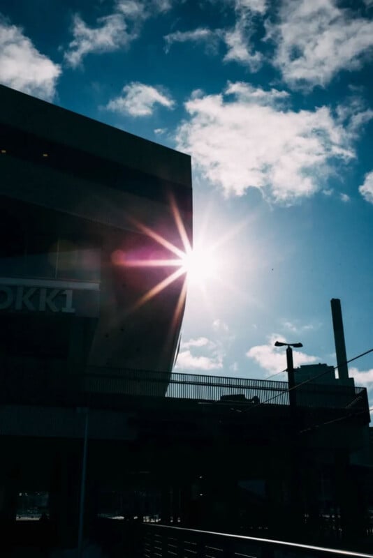 The sun shines brightly behind the corner of a modern building, creating a starburst effect in a partly cloudy blue sky. Shadows and silhouettes of structures and a lamppost are visible in the foreground.