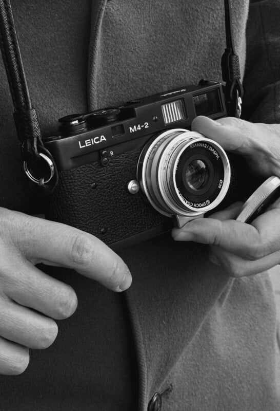 A person holding a vintage Leica M4-2 camera, adjusting the lens with one hand. The camera hangs from a strap around their neck; the image is in black and white.