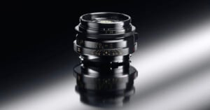 A close-up of a black camera lens with gold markings, standing upright on a glossy, reflective surface against a dark background.
