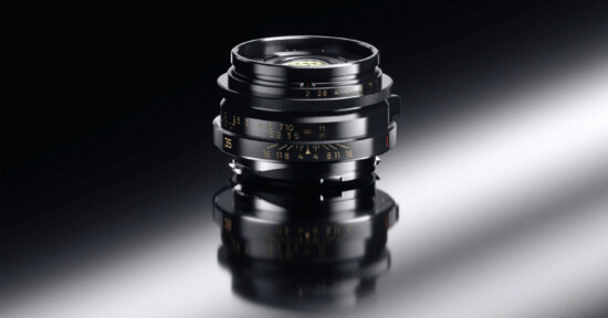 A close-up of a black camera lens with gold markings, standing upright on a glossy, reflective surface against a dark background.