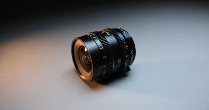 A close-up of a professional camera lens with focus and aperture rings, resting on a smooth, neutral-colored surface under soft lighting.