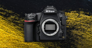 A Nikon D850 DSLR camera body without a lens is displayed against a textured black and yellow background. The camera’s sensor is visible in the center where the lens would attach.