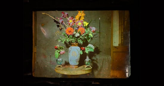 A vintage photograph of a blue vase with colorful flowers on a small round table, flanked by two glass containers holding single flowers, set against a textured wall.