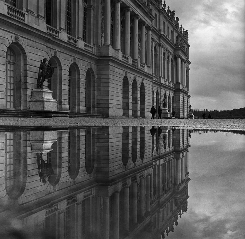 A grand, ornate building with tall columns and arched windows is reflected in a large puddle on the ground; people and a statue are visible near the entrance under a cloudy sky.