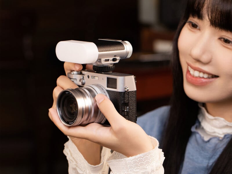A smiling woman holds a vintage-style camera with both hands, aiming it forward. The camera has a large lens and an external attachment on top. The woman is indoors and wearing a blue top with a white lace collar.