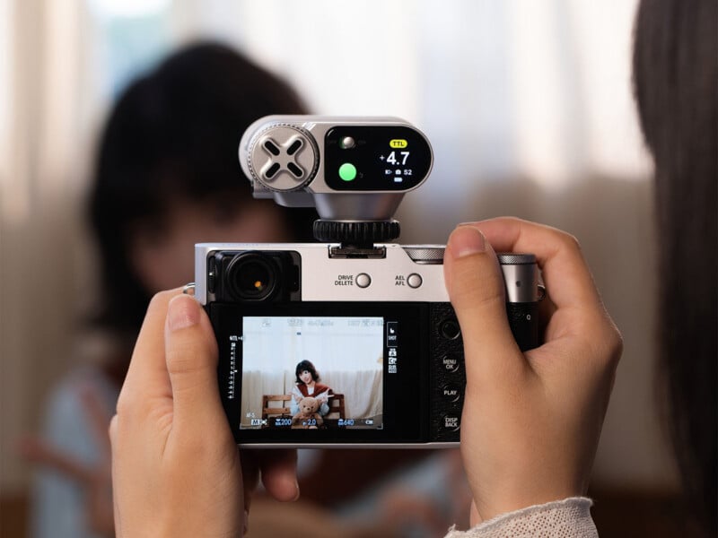 Close-up of hands holding a digital camera, focusing on a screen showing a young girl sitting on a sofa. An external device with a digital display is mounted on the camera. The background is softly blurred.