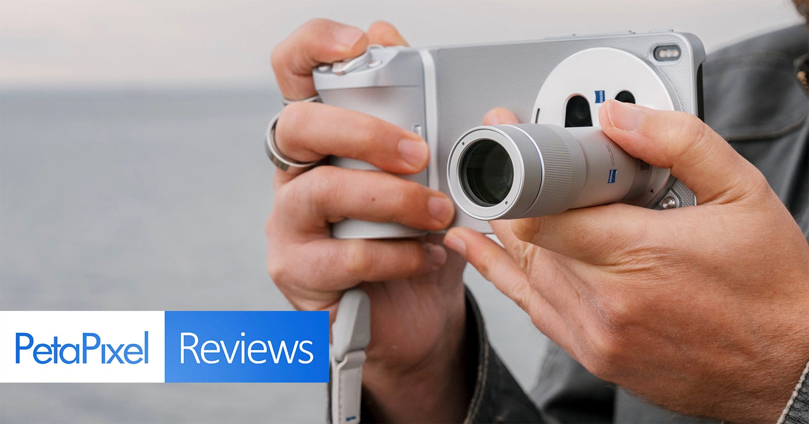 A person holding and using a silver camera with both hands near a body of water. The image features the "PetaPixel Reviews" logo in the bottom left corner.
